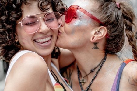 Two people in rainbow-themed outfits embrace and smile; one kisses the other on the cheek at a pride event. Both wear sunglasses and appear joyful, with pride flags and festive atmosphere in the background.