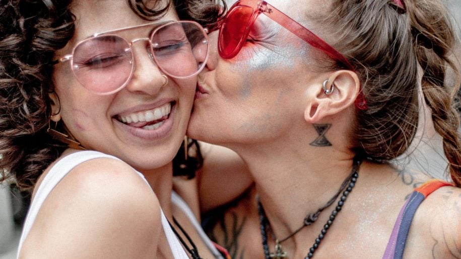 Two people in rainbow-themed outfits embrace and smile; one kisses the other on the cheek at a pride event. Both wear sunglasses and appear joyful, with pride flags and festive atmosphere in the background.