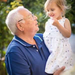 An elderly man with white hair and glasses holds a smiling young girl wearing a white dress. They look at each other affectionately, standing outdoors with greenery in the background.