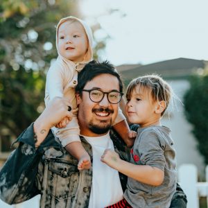 A smiling man with glasses holds a baby on his shoulder and hugs a young child, standing outside by a white picket fence with trees and a house in the background.