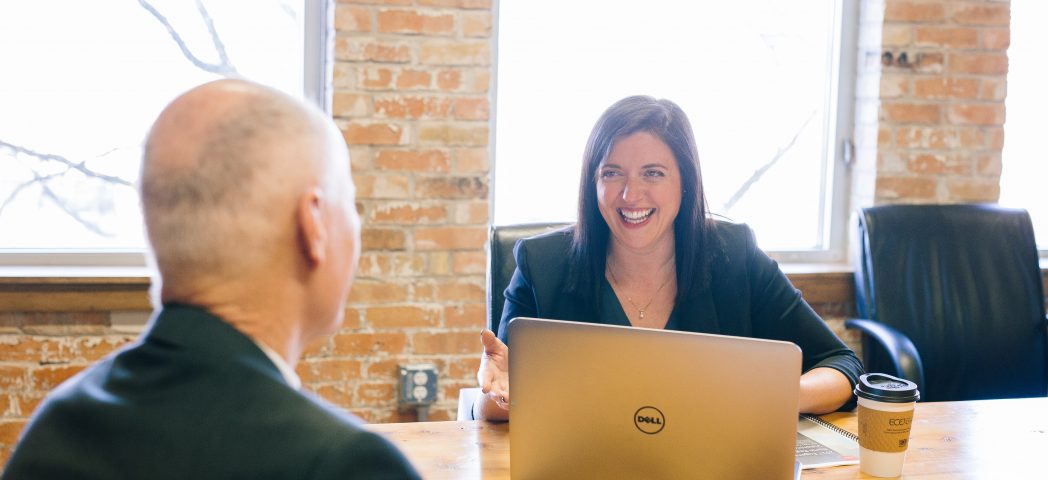 A woman smiles whilst talking to a man across a table in an office. She sits behind a laptop, holding a pen. Both are dressed in business attire. Coffee cups are on the table. Large windows and brick walls are in the background.